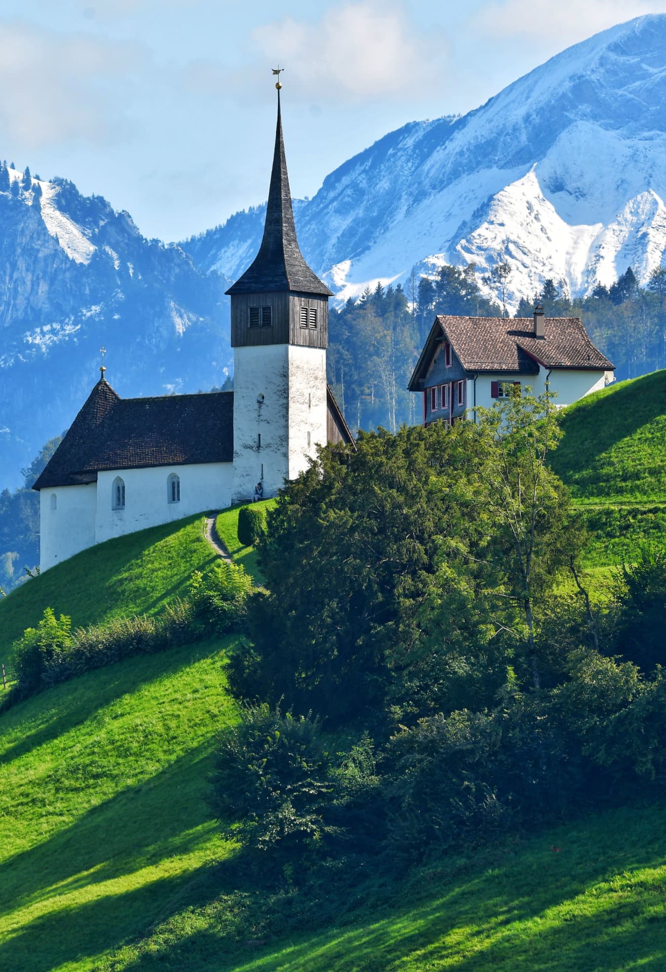 White chapel on a green hillside beneath snowy alpine peaks